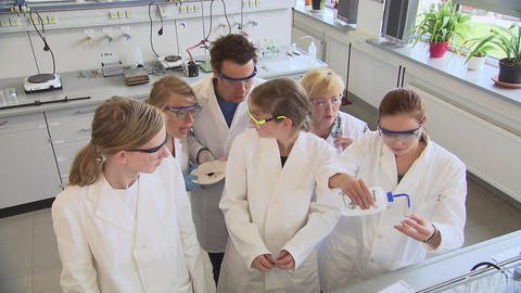 Sechs Personen stehen in weißen Kitteln in einem Labor und experimentieren.