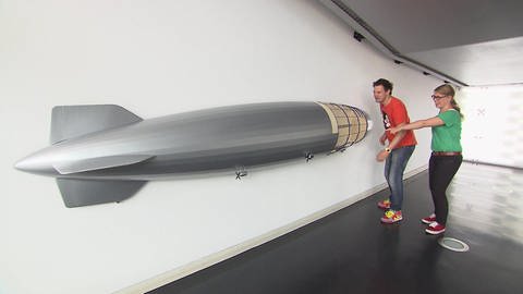 Ein Mann und eine Frau stehen in einer Halle vor dem Modell eines Zeppelins. 