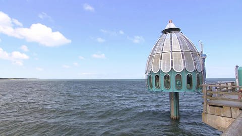 Ein glockenförmiges, kleines, türkises Haus ragt auf einer Stelze über dem Meer.