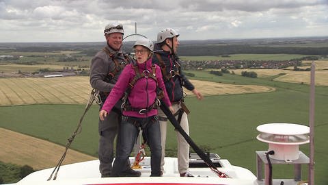 Drei Personen stehen mit Klettergurten und Helmen gesichert auf einem Windrad.
