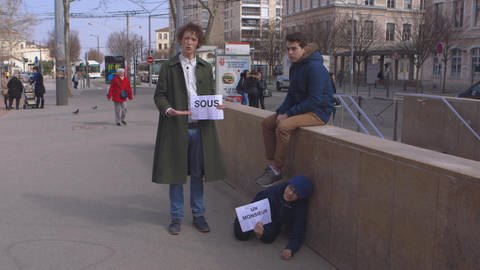 Ein junger Mann steht in einer Stadt und hält ein Schild, darauf steht "sous". Daneben kauert ein Junge unter einem Jungen auf einer Mauer und hält ein Schild mit "un monsieur". 